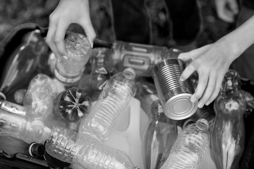 Black and white photograph showing two hands reaching into a collection of discarded recyclable bottles and cans. Several clear plastic bottles with visible ridges and caps, along with a few metal cans, are scattered in a container or on a surface. The bottles vary in size and shape, some with labels or markings, while the cans are plain with smooth metallic finishes. The scene appears to be outdoors, with a blurred background suggesting natural surroundings or a waste collection area. The lighting highlights the textures of the plastic and metal surfaces, emphasizing the differing materials. This image illustrates waste separation or recycling efforts, relevant to rubbish removal services provided by Office Clearance Soho in London, often serving areas within postcode districts like Soho or central London neighborhoods.