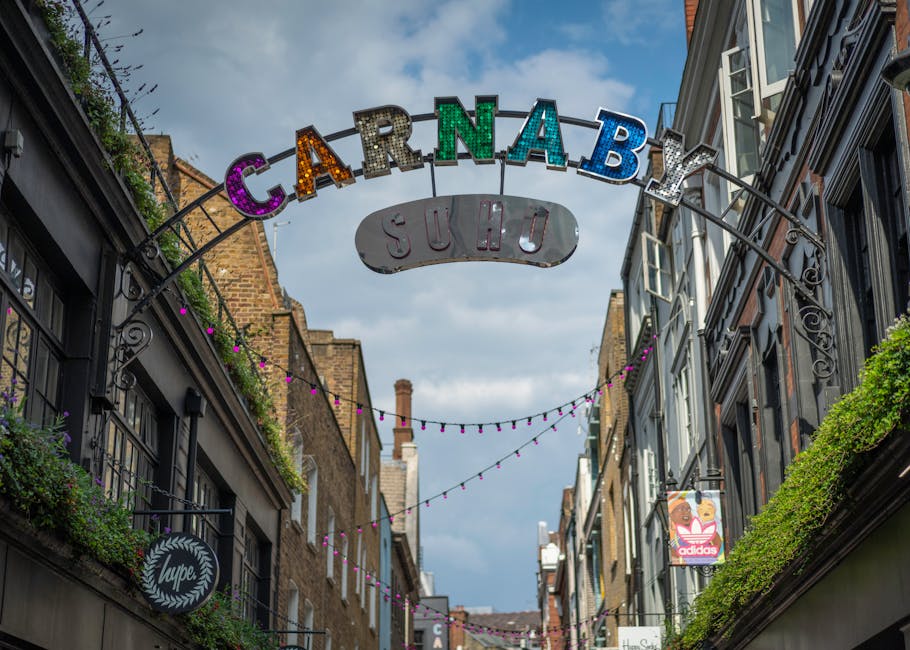 A street scene in Soho featuring an overhead sign with the word 'CARNABY' in large, colorful, letter-shaped lights, with the smaller sign below reading 'SUHO' in white letters on a grey background. Th