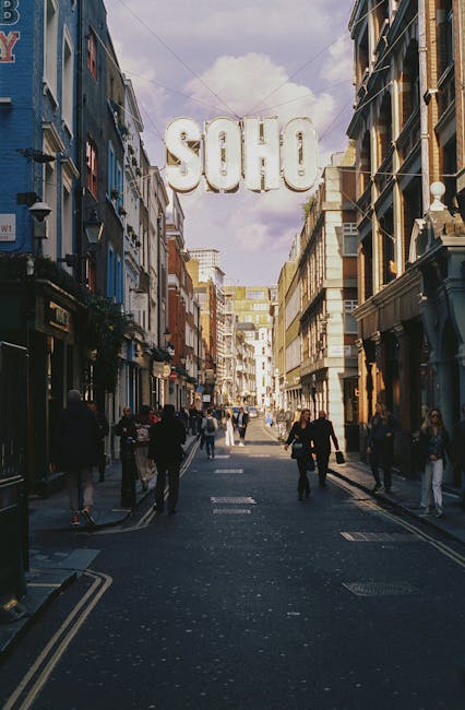 A busy street scene in central London featuring a large, white, illuminated sign spelling out 'SOHO' suspended above the roadway. The street is lined with multi-storey buildings on both sides, showcas